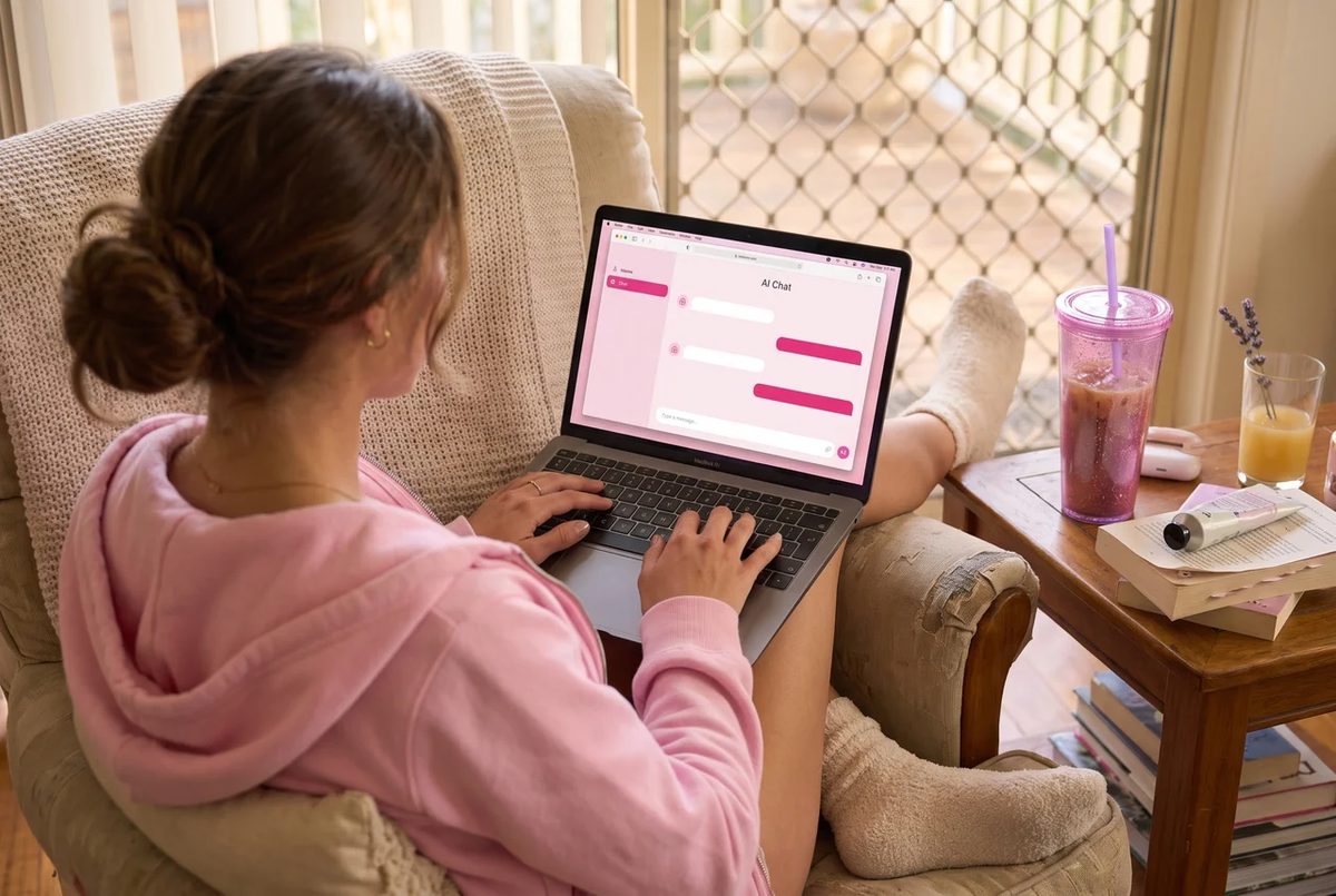 Over-the-shoulder view of a woman in a pink hoodie typing on a laptop in an armchair.
